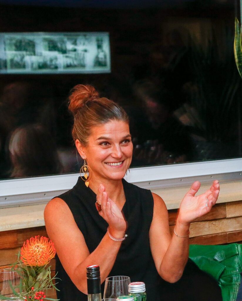 Eine lächelnde Frau im dunklen Oberteil klatscht in die Hände; im Hintergrund spiegelt sich eine nächtliche Szene im Fenster