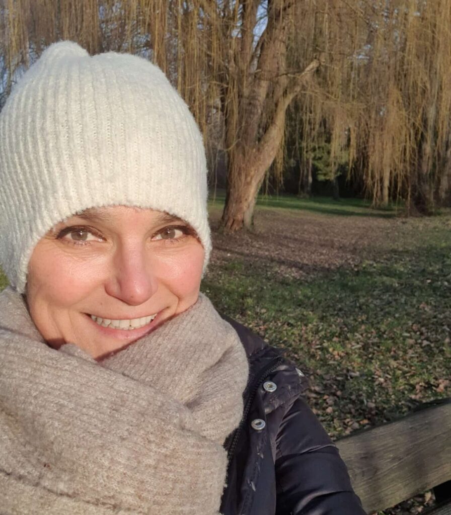 Eine Frau mit einer hellen Strickmütze und einem Schal macht ein Selfie im Freien vor winterlichen Bäumen