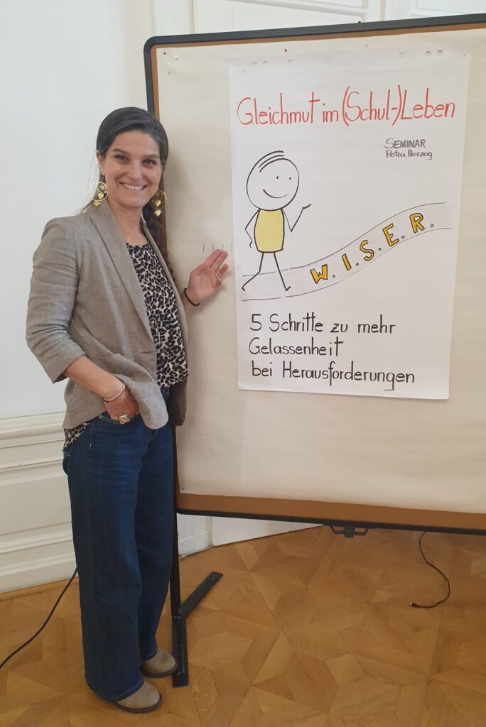 Eine Frau steht neben einem beschrifteten Plakat, auf dem eine Strichzeichnung und der Text „5 Schritte zu mehr Gelassenheit bei Herausforderungen“ zu sehen sind