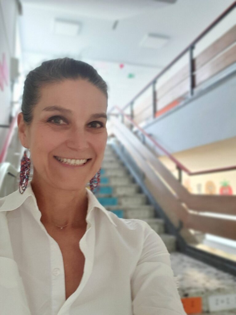 Eine lächelnde Frau blickt in die Kamera; im Hintergrund ist ein helles Treppenhaus zu sehen