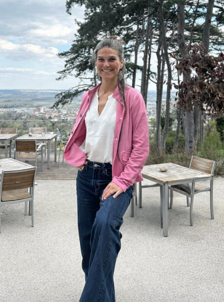Eine lächelnde Frau im weißen Top, einer rosafarbenen Jacke und Jeans steht im Außenbereich eines Cafés