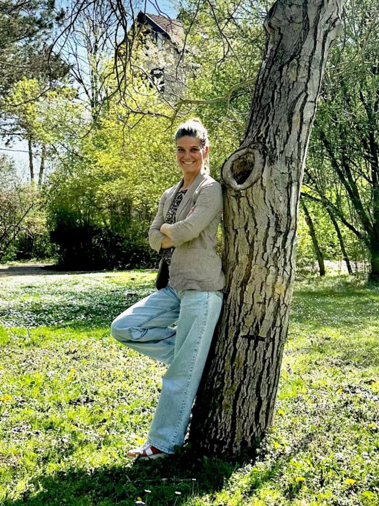 Eine Frau in heller Kleidung lehnt entspannt an einem großen Baumstamm im Park