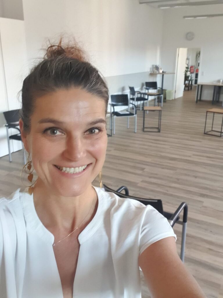 Eine lächelnde Frau mit hochgesteckten Haaren macht ein Selfie in einem hellen, leeren Raum mit Holzboden
